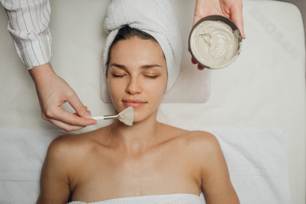 Hands of a Caucasian cosmetologist woman applying a facial mask on a beautiful young Caucasian woman's face lying on the massaging bed.